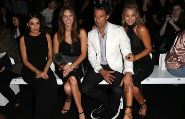 Four people seated at a fashion show, smiling for the camera, with a crowd in the background.
