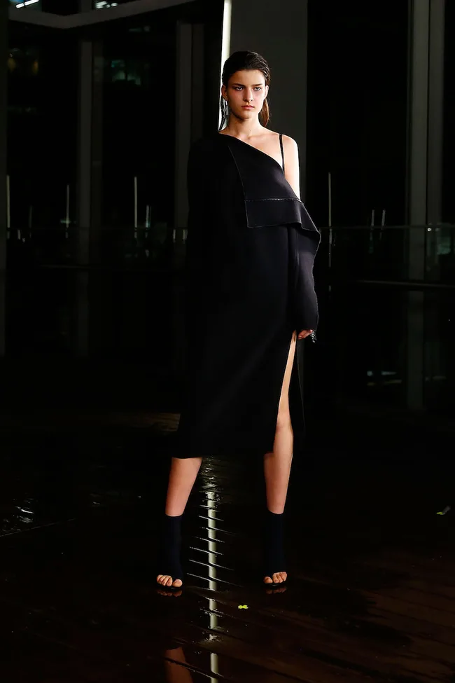 Model wearing a black off-shoulder dress and heels on a wet runway, with a dark background.