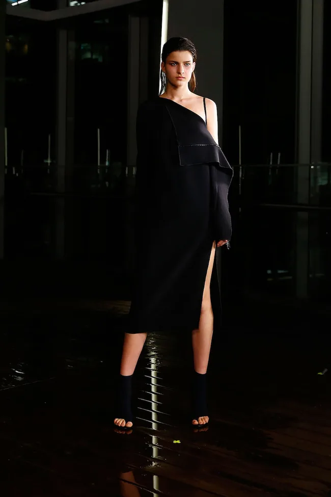 Model wearing a black off-shoulder dress and heels on a wet runway, with a dark background.