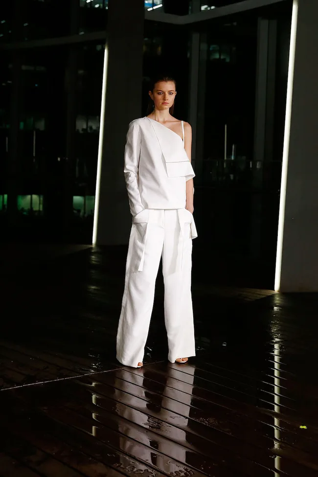 Model in an asymmetrical white outfit stands on a wet, dark runway at night.