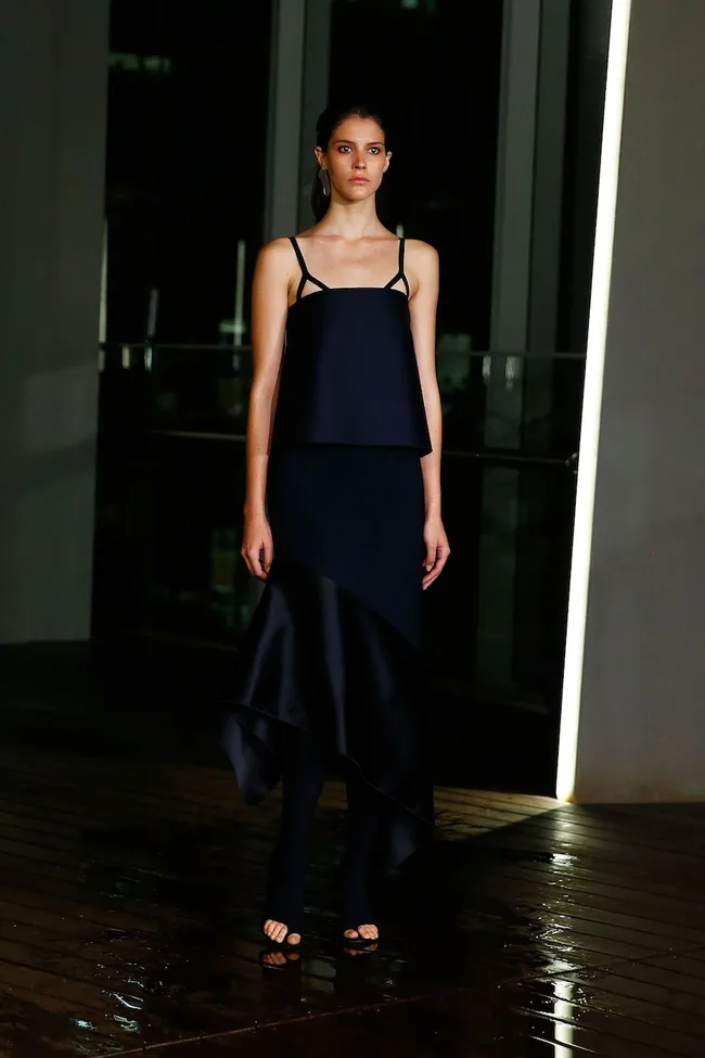 A model in a sleek black dress with asymmetrical hem stands indoors on a wet, wooden floor.