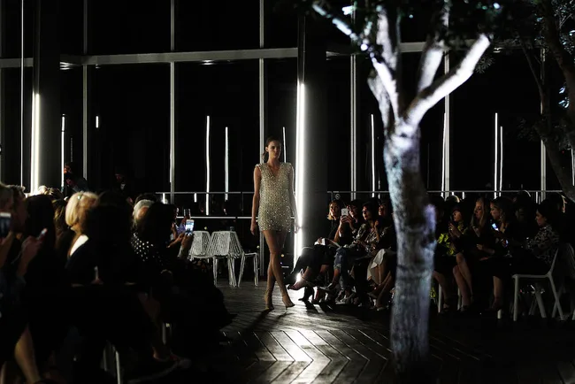 Model walking down a dimly lit runway, surrounded by an audience in a modern venue with trees.