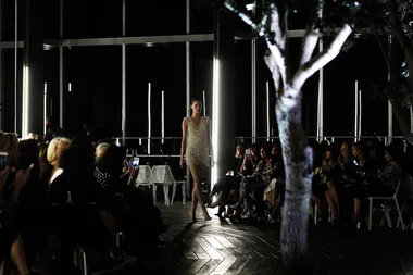 Model walking down a dimly lit runway, surrounded by an audience in a modern venue with trees.