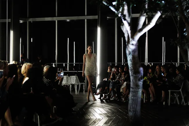 Model walking down a dimly lit runway, surrounded by an audience in a modern venue with trees.