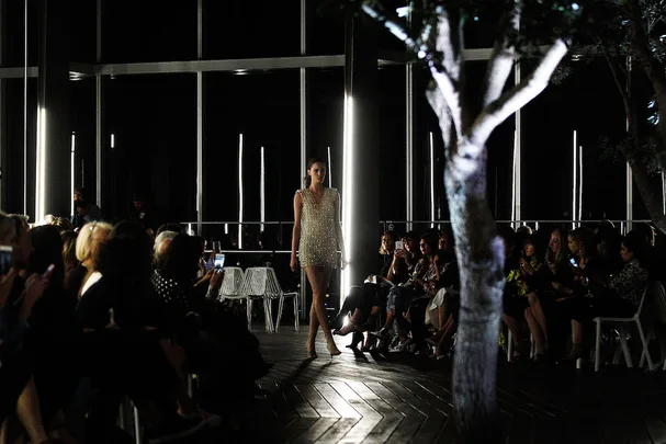 Model walking down a dimly lit runway, surrounded by an audience in a modern venue with trees.