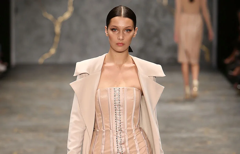 A model in a beige strapless dress and coat walks down the runway with a neutral expression.