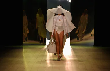Model in white lace outfit and oversized hat walking down runway with matching handbag.
