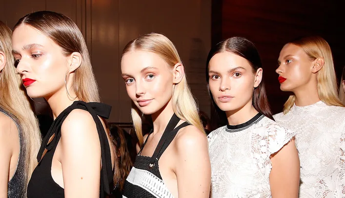 Backstage at a fashion show with models in sleek hairstyles and stylish outfits, standing in line.