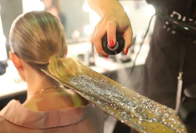 A hairstylist sprays glitter on a woman's sleek, tied-back hair in a salon setting.