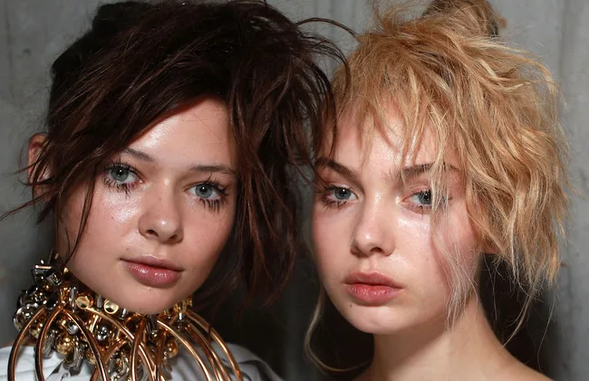 Two models with intricate hairstyles and shimmering makeup, posing closely together against a plain backdrop.