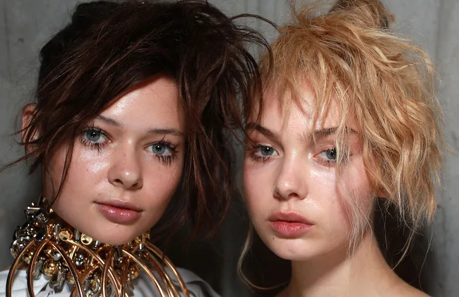 Two models with intricate hairstyles and shimmering makeup, posing closely together against a plain backdrop.