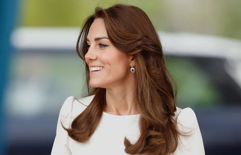 A woman with long brown hair, smiling and wearing a white top and dark gemstone earrings.
