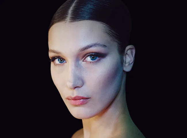 A close-up of a woman with dark, slicked-back hair against a black background, showcasing her striking blue eyes and neutral expression.