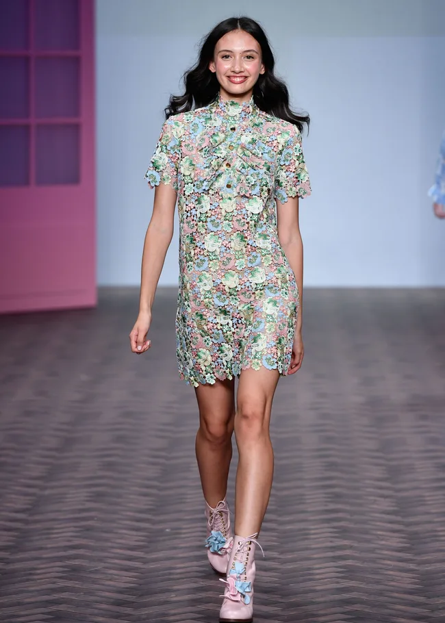 Model in a floral lace dress and pastel shoes walks the runway with a smile, pink door in the background.