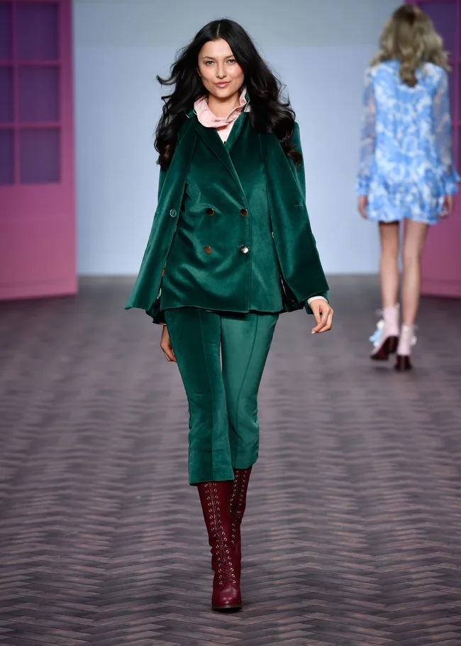 Model in green velvet suit and maroon boots walks runway, with another model in blue behind.