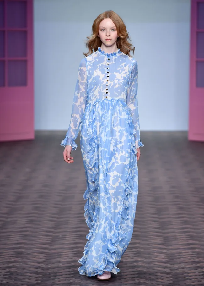 Model in long blue and white patterned dress walks on runway, with pink doors in the background.