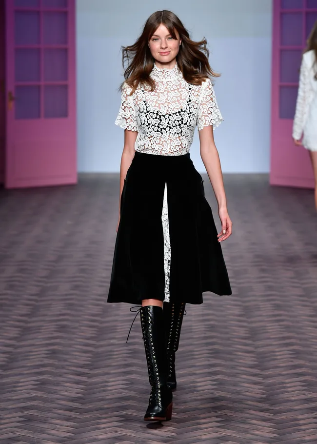 Model in a white lace top and black skirt walking on a runway with pink doors in the background.