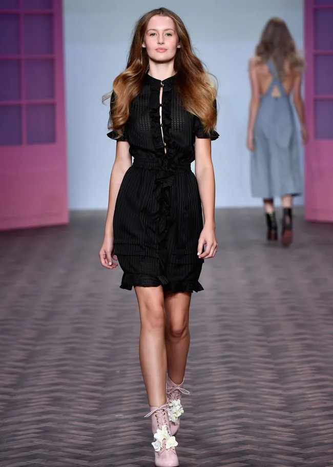 Model wearing a black dress and pink floral boots walks on a fashion runway, with another model in the background.