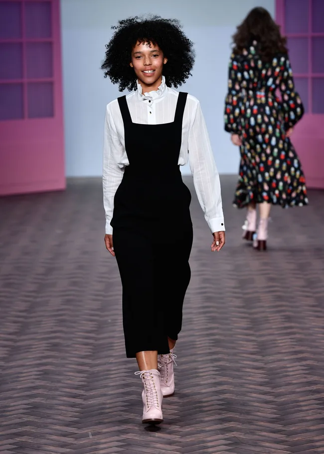 Model walking in fashion show wearing a white blouse and black pinafore dress with pink lace-up boots.