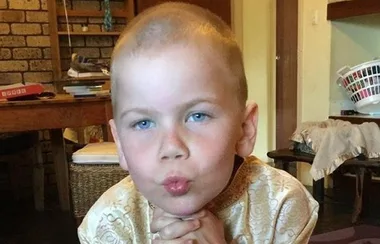 Young child with short hair, wearing a patterned shirt, rests chin on hands and puckers lips in an indoors setting.