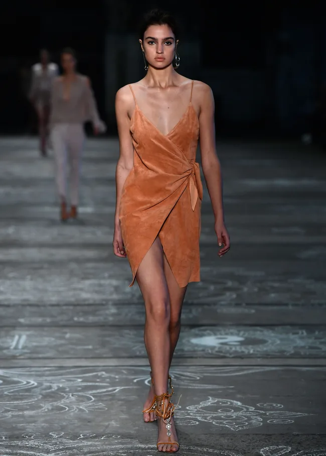 Model in a brown wrap dress walks on a runway, wearing dangling earrings and strappy sandals.