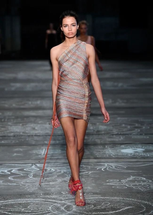 Model in a colorful one-shoulder dress and red heels walking on a runway with a patterned floor background.
