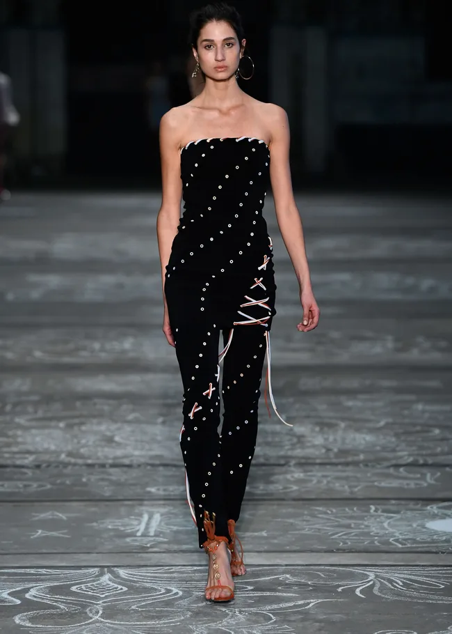 Model in a strapless black dress with eyelet details walks on a patterned runway, wearing hoop earrings and sandals.