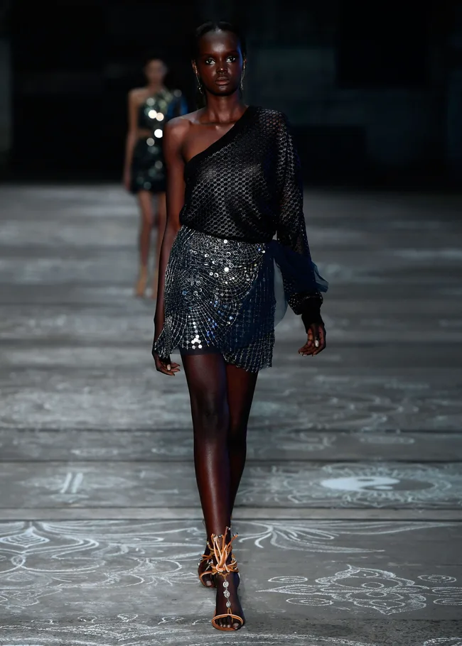 Model walks runway in a dark, sequined one-shoulder outfit with intricate patterns and orange strappy heels.