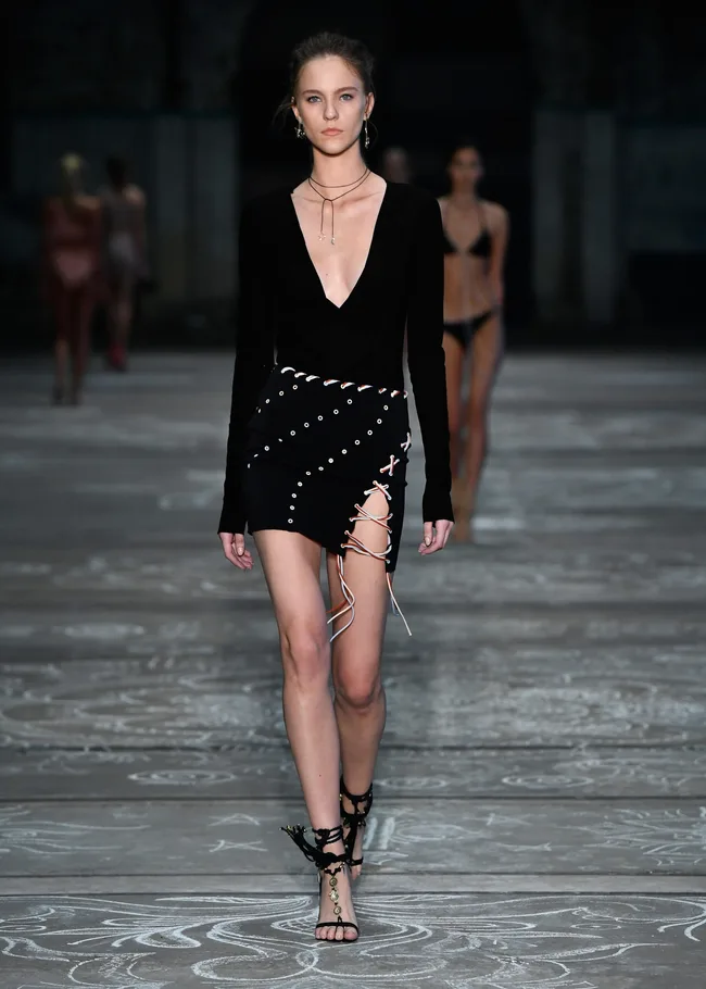 Model in black dress with eyelet and lace details walks runway, wearing strappy sandals, with others in background.