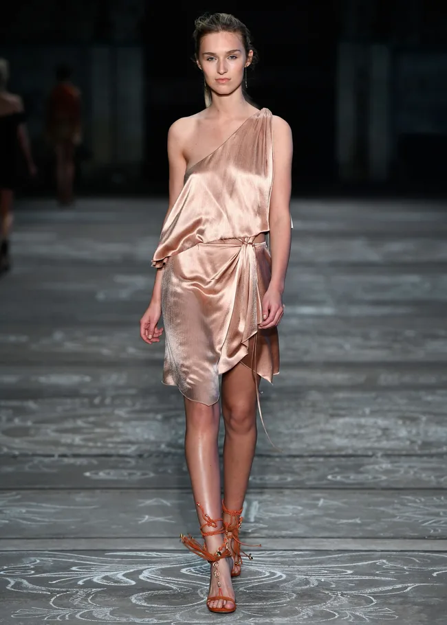 Model in a silky, one-shoulder dress and orange sandals walking on a runway.