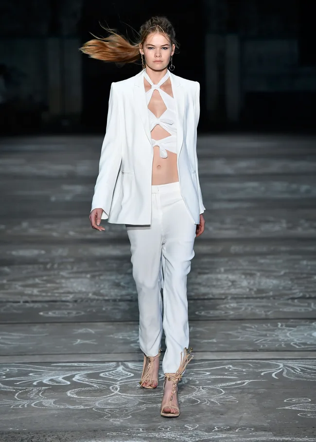 Model walks in white cut-out suit and sandals on a runway, hair styled back.