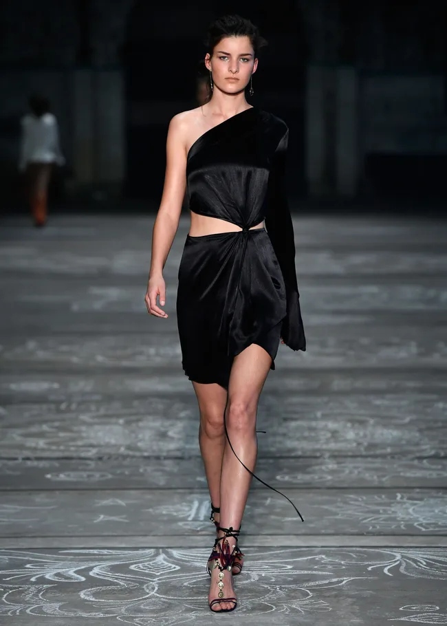 Model in a black asymmetrical dress and sandals walking on a runway at a fashion show.