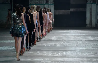Runway models in stylish outfits walking in line during a fashion show in an industrial-style venue.