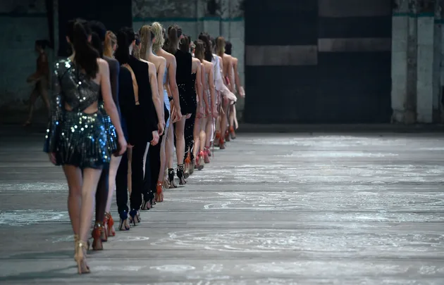 Runway models in stylish outfits walking in line during a fashion show in an industrial-style venue.