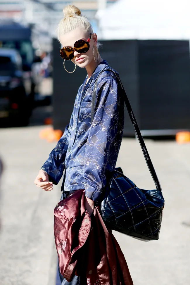 A person with blonde hair in a bun, wearing large sunglasses, a blue patterned shirt, and a quilted bag.