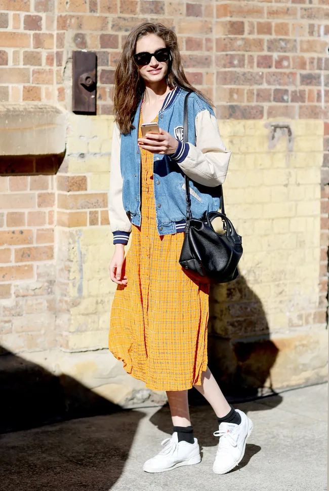 Woman in denim jacket, orange dress, and sneakers holds a phone, smiling against a brick wall backdrop.