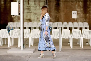 Woman in patterned blue dress walks near white chairs, holding a navy handbag, on a city street.