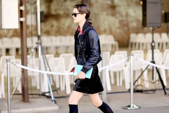 A person in a black outfit and sunglasses walks past rows of white chairs, holding a colorful book.