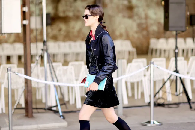 A person in a black outfit and sunglasses walks past rows of white chairs, holding a colorful book.