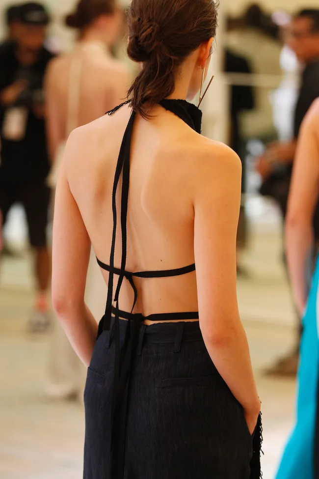 Back view of a person in a black halter top and dark pants, with hair in a bun, hands in pockets, in a blurred background.