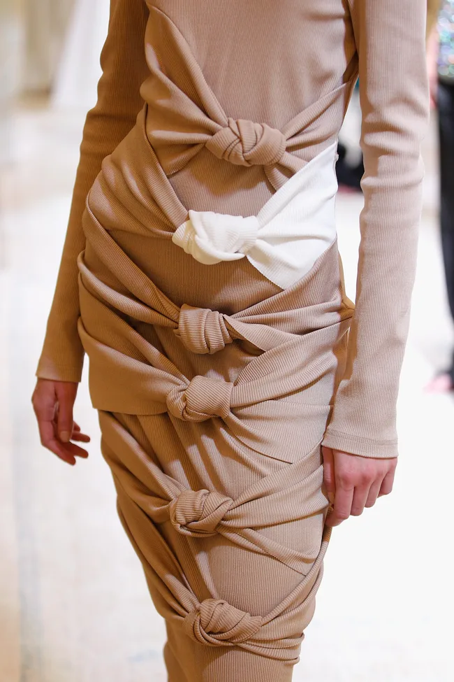 Dress with tan fabric featuring knotted details and one white knot near the waist, worn by a model on a runway.