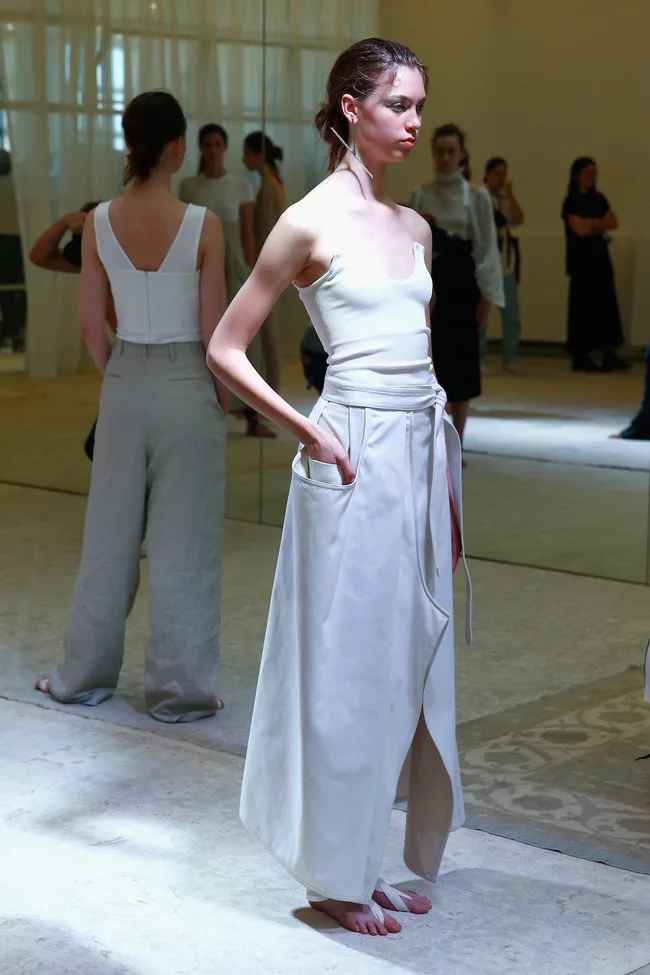 Model in a white tank top and long skirt with pockets, barefoot, at a fashion event with others in the background.
