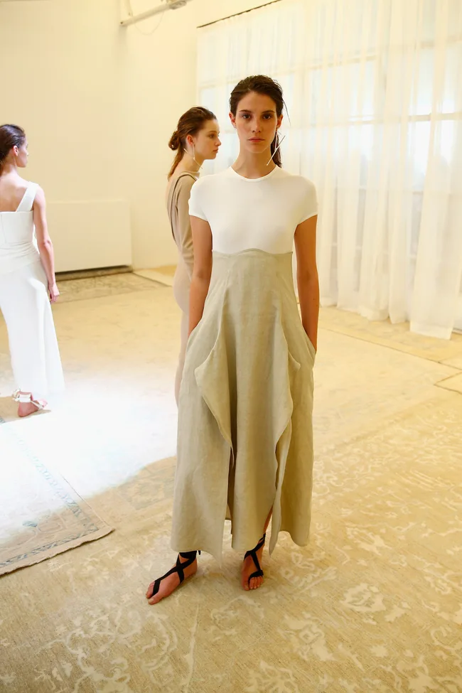 Models showcase elegant, neutral-toned outfits with draped fabrics on a patterned carpet.