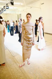 Model in a beige dress with knot details at a fashion show, standing among other models in varied outfits.