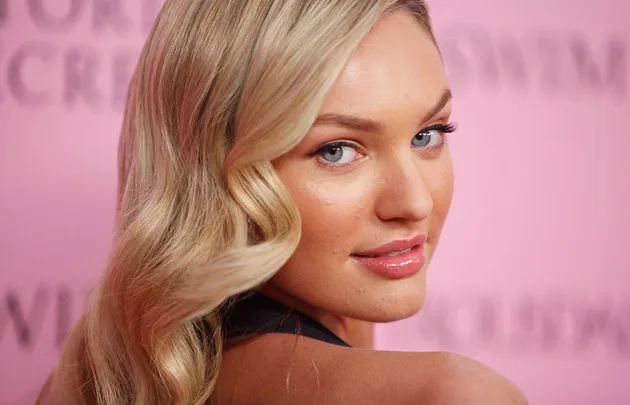 Blonde woman with curled hair poses against a pink background, displaying a slight smile while looking over her shoulder.
