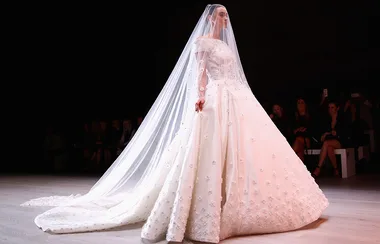 Model in an elegant white wedding gown with a long veil walks on a runway, surrounded by an audience in dim lighting.