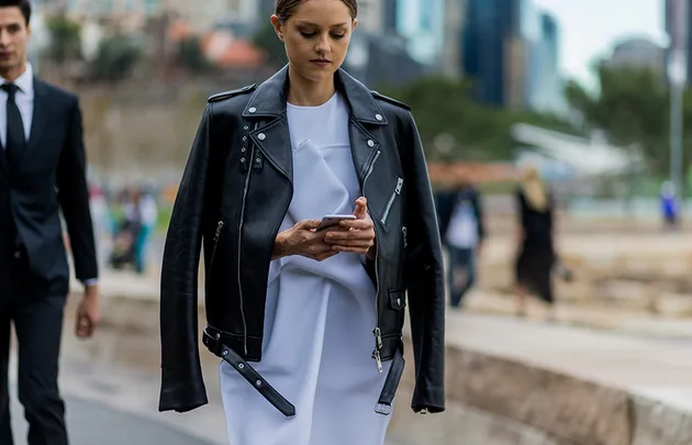 Woman in a black leather jacket over a white dress, looking at her phone while walking in an urban setting.