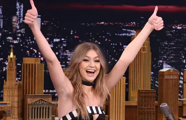 A woman smiling with both thumbs up on a talk show set, with a city skyline backdrop at night.