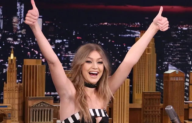 A woman smiling with both thumbs up on a talk show set, with a city skyline backdrop at night.
