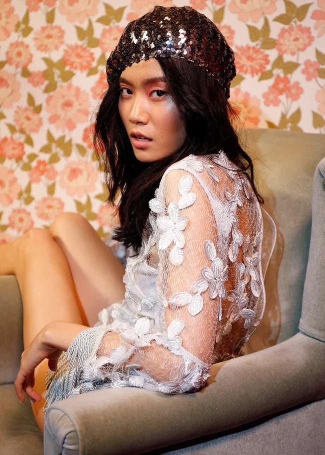 Woman in a floral lace dress and sequined beret sitting against a floral wallpaper backdrop.